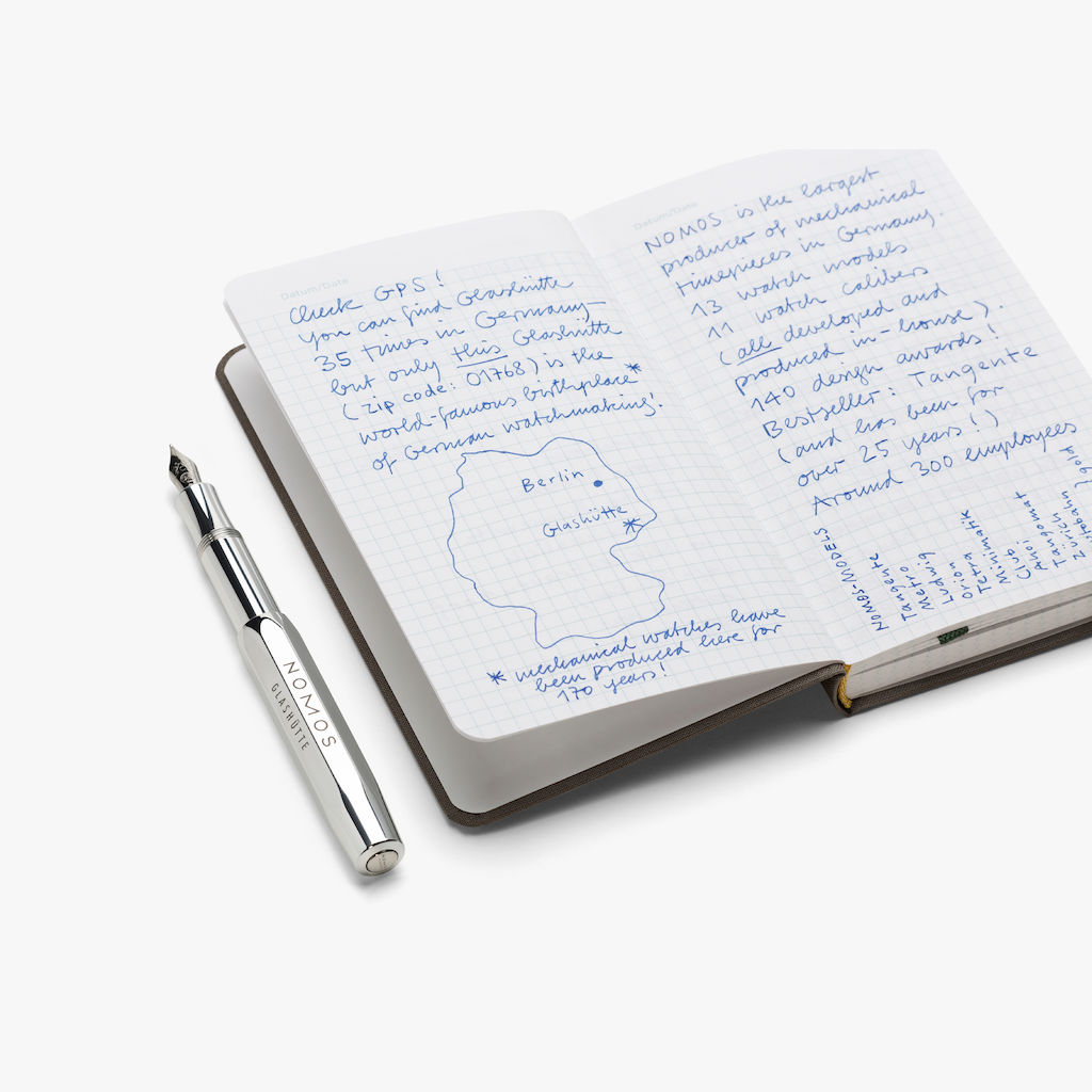 Hard cover notebook with a gray linen binding and matt silver NOMOS embossing, size: 19.5 by 12.5 centimeters. With 192 bound pages from graph paper (90 g/qm), an olive-green bookmark ribbon, an elastic closure, and an inner pocket on the back cover.