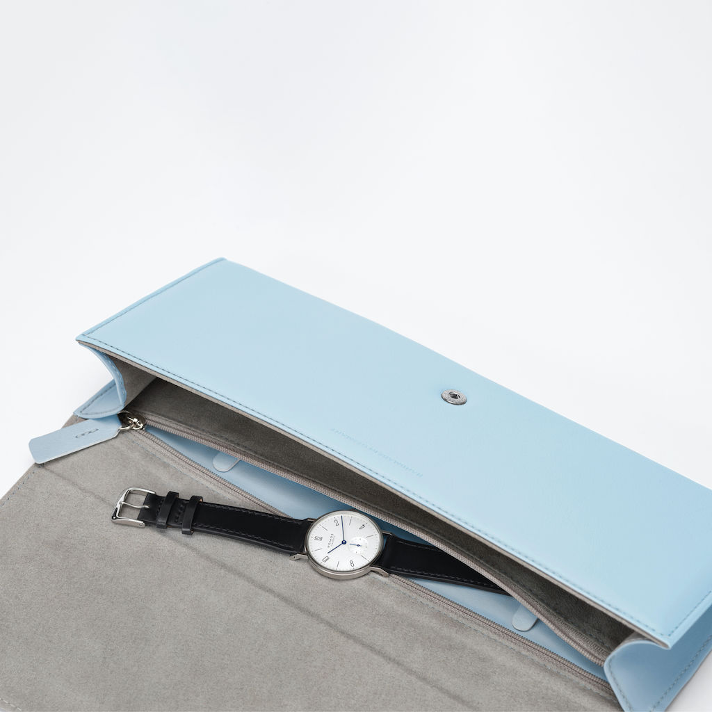 Clutch NOMOS in pelle bovina, conciata senza uso di cromo, resistente alla luce e con interno rivestito. Dimensioni: 30 x 12,5 cm. Chiusura: bottone a pressione cucito internamente. Scomparto separato per orologio con cerniera.